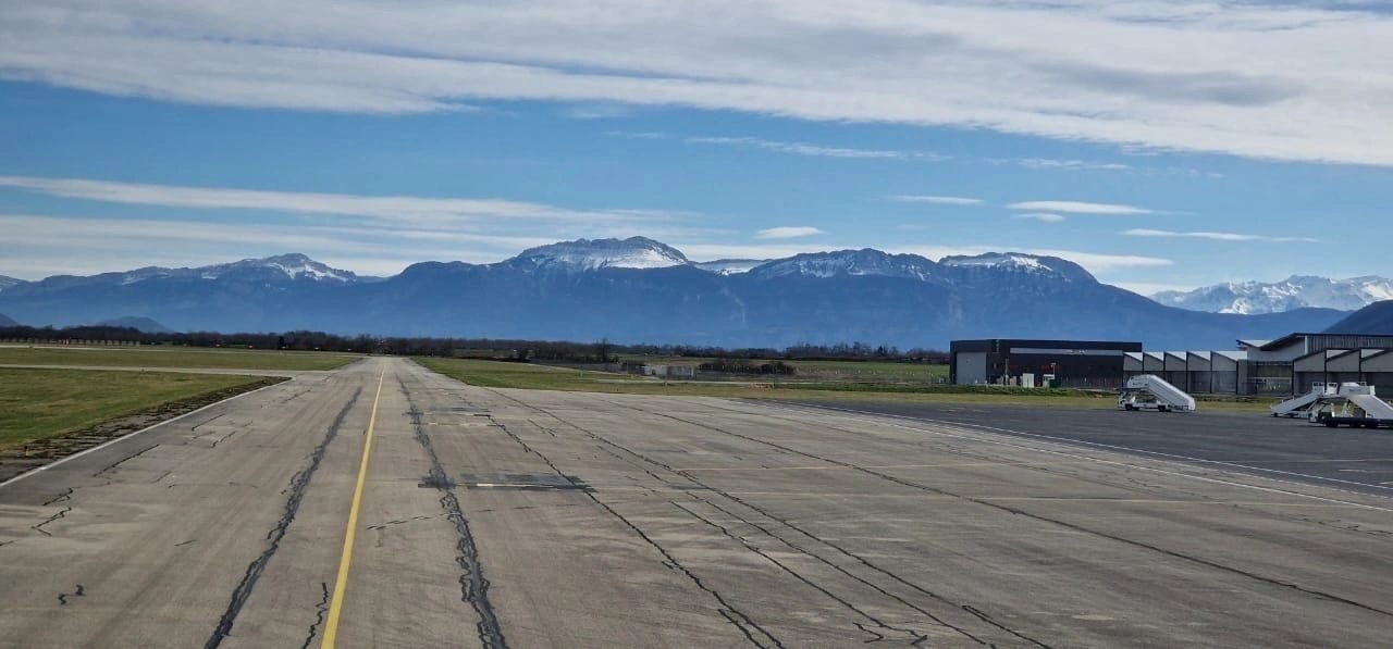 A runway leading towards distant mountains, with aircraft parked on the apron
